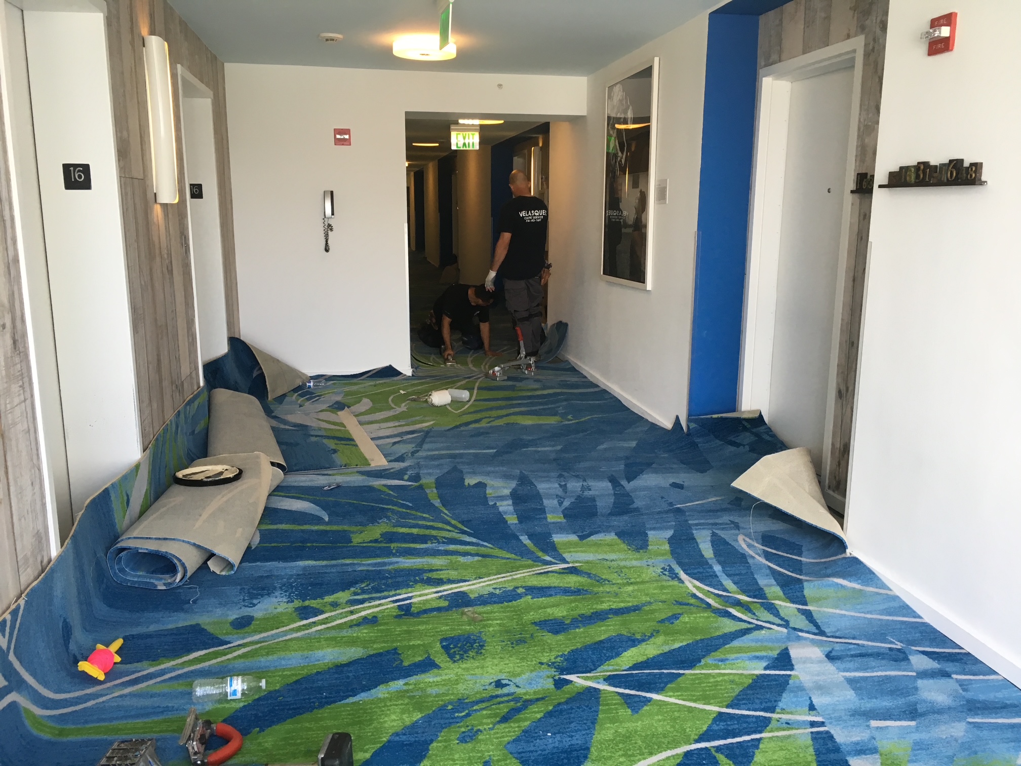 Flooring contractors installing a custom blue and green tropical leaf patterned commercial carpet in a hotel corridor.