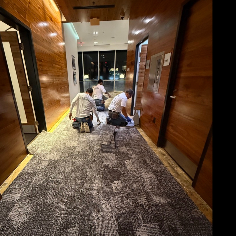 Professional installers laying down multi-toned gray patterned commercial carpet tiles in a wood-paneled hallway.